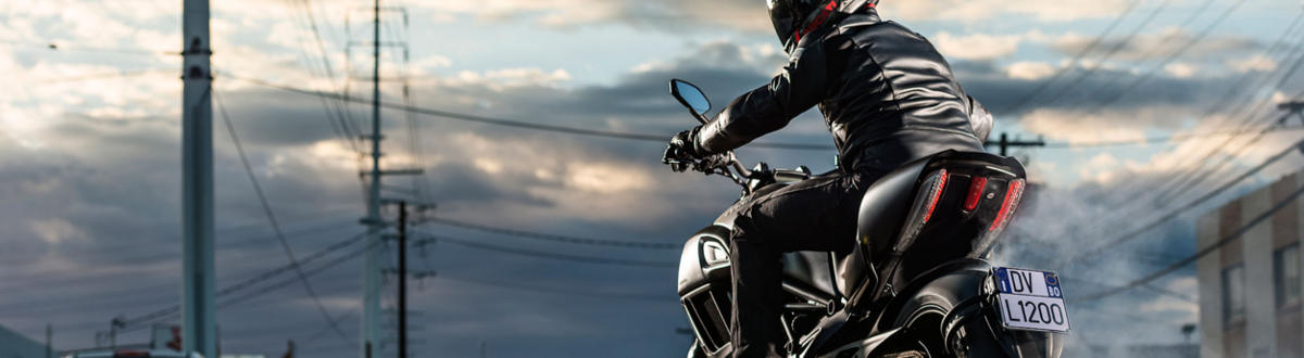A guy with a black jacket on riding a black motorcucle looking backwards at the camera